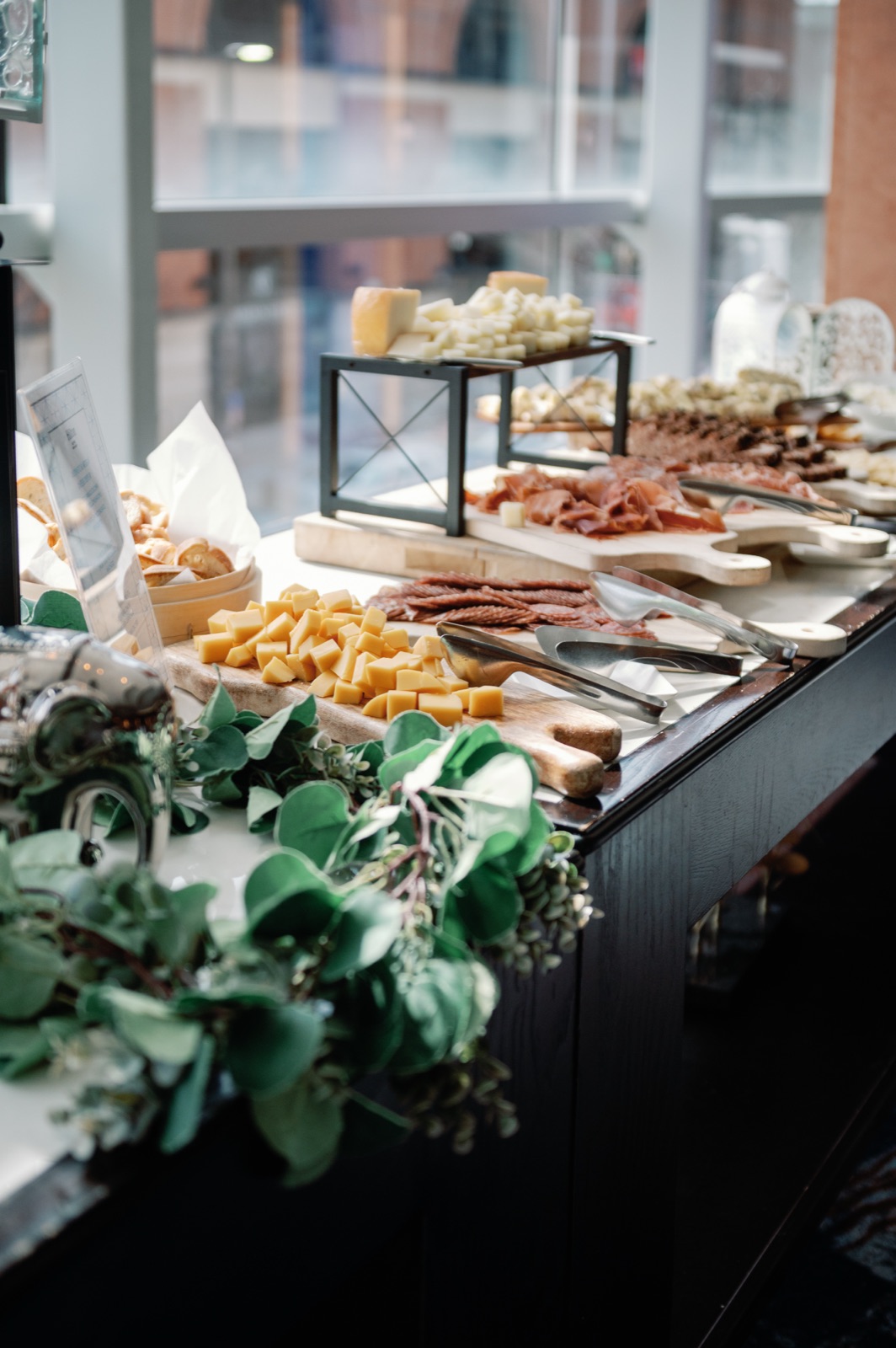 Grazing display of cheeses and charcuterie on wooden boards, edged with eucalyptus, beside a sunlit window