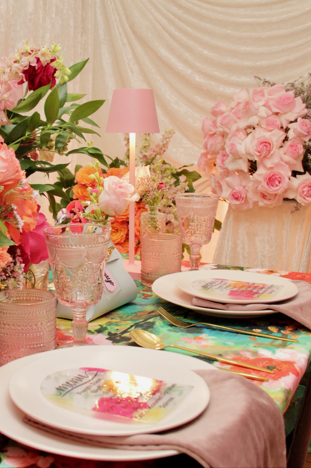 Styled table with pink roses, a blush lamp, patterned linens, and custom place cards
